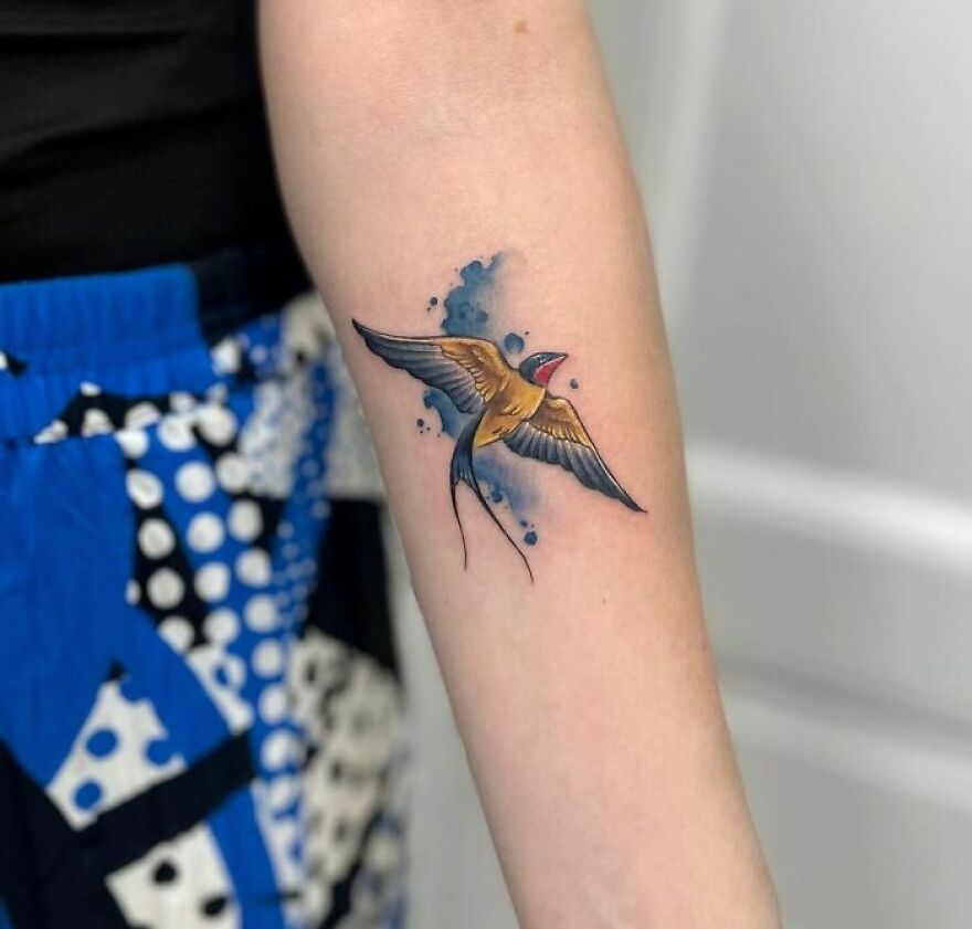 Watercolor tattoo of a vibrant swallow on forearm, featuring blue splashes, showcasing artistic ink design.