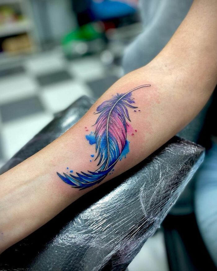 Watercolor tattoo of a colorful feather on a forearm, blending shades of blue and pink for inkspiration.