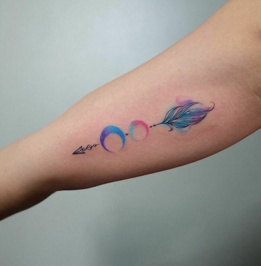 Watercolor tattoo with crescent moons and a feather on an arm, showcasing vibrant hues.