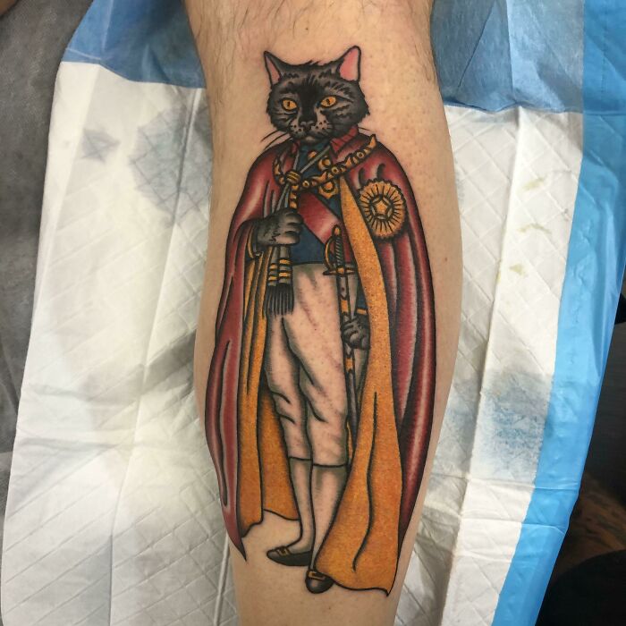 Adorable pet tattoo of a royal cat wearing a cape on a person's calf.