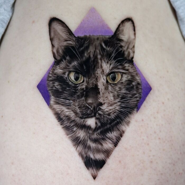 Pet tattoo of a cat's face with a purple diamond background on skin.