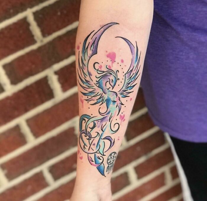 Watercolor tattoo on forearm featuring a vibrant, abstract phoenix design with blue and pink hues for inkspiration.