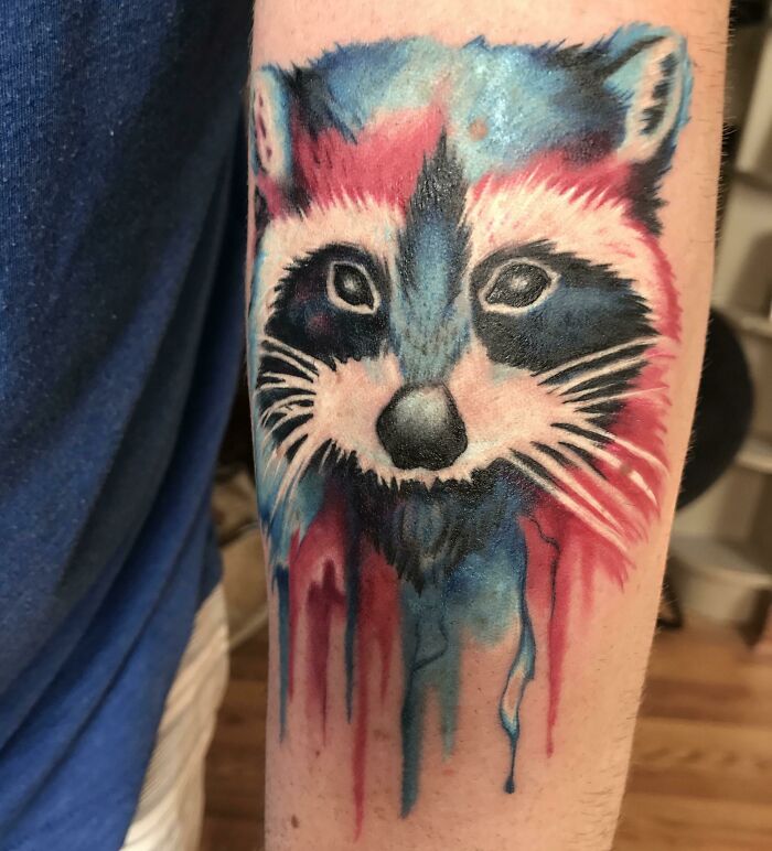 Watercolor tattoo of a raccoon with vibrant blue and red hues dripping down the arm.