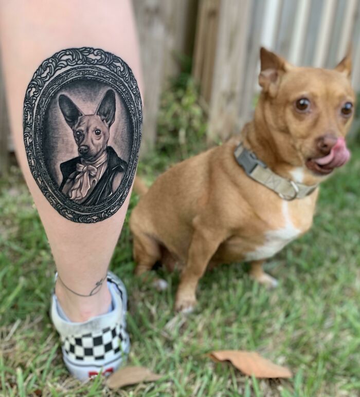 A pet tattoo on a person's leg, depicting a dressed-up dog in an ornate frame, with a matching dog beside them.