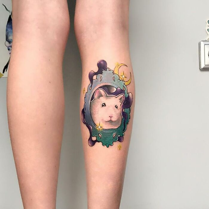 Pet tattoo of a white animal in a colorful, decorative frame on a person's calf.