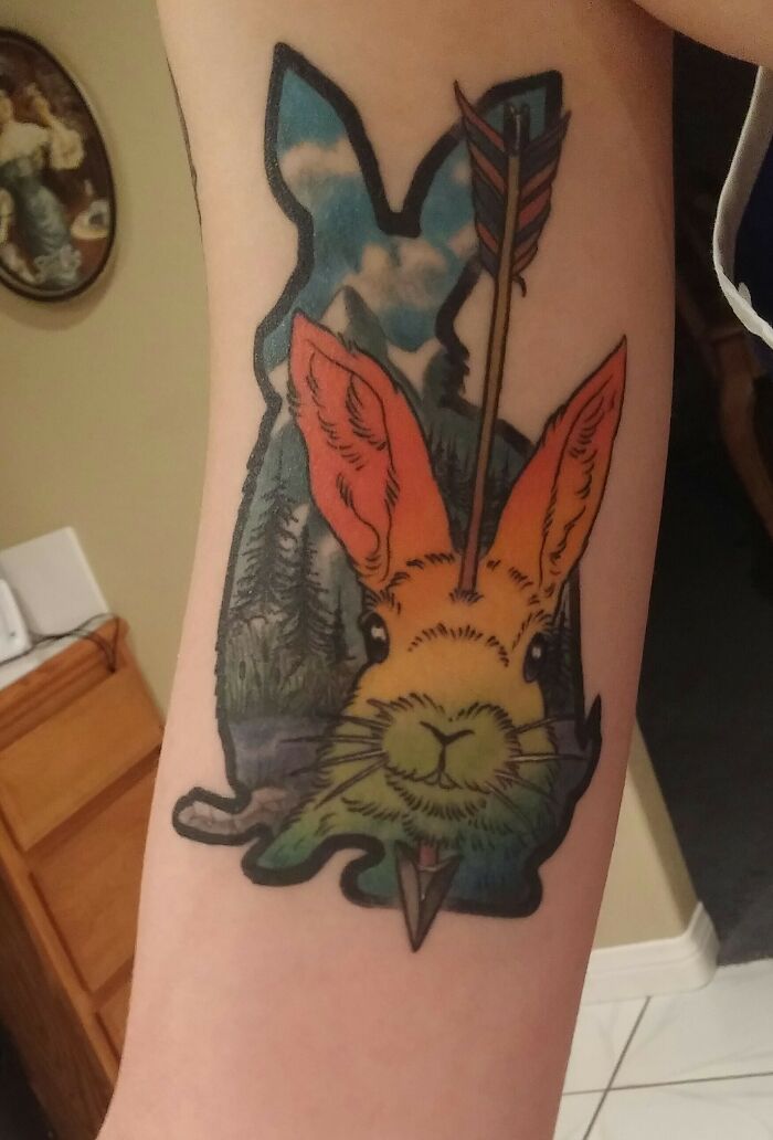 Colorful pet tattoo of a rabbit with an arrow through its ear, set against a forest landscape, celebrating furry friends.