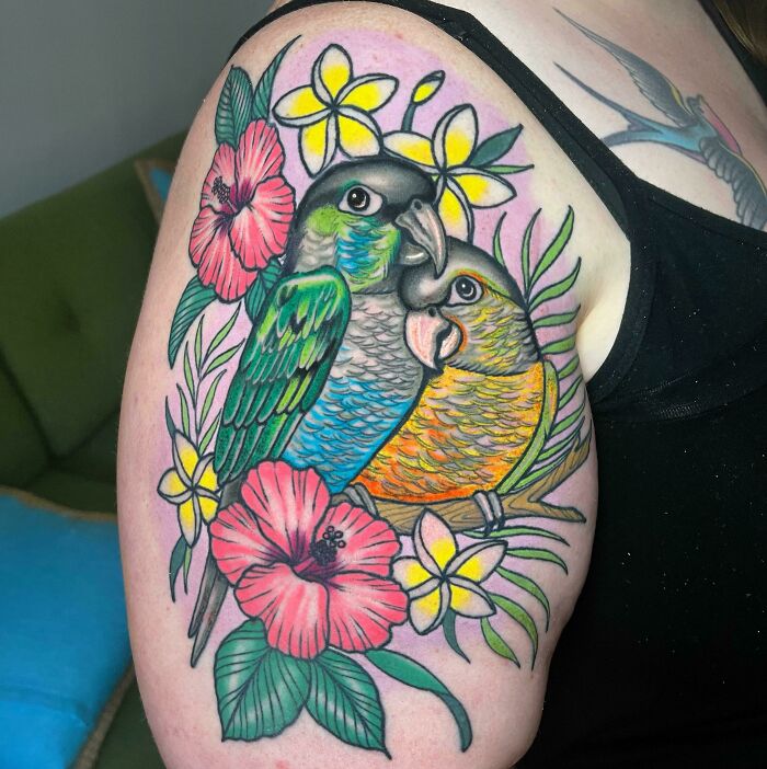 Colorful tattoo of two pet parrots surrounded by flowers on an arm.