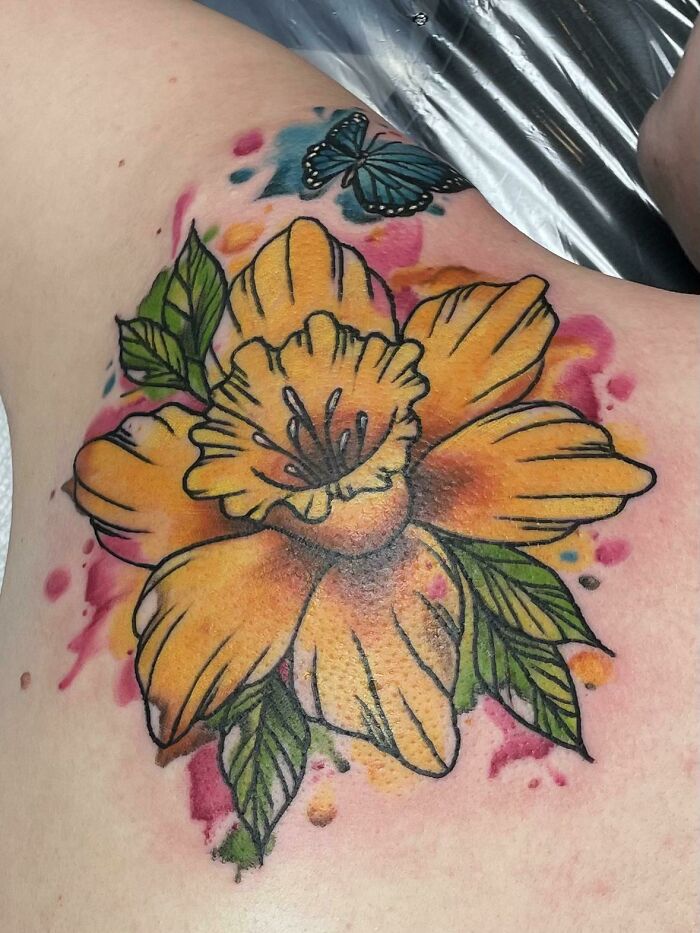 Watercolor tattoo of a yellow flower with leaves and a blue butterfly on skin, showcasing ethereal artistry.