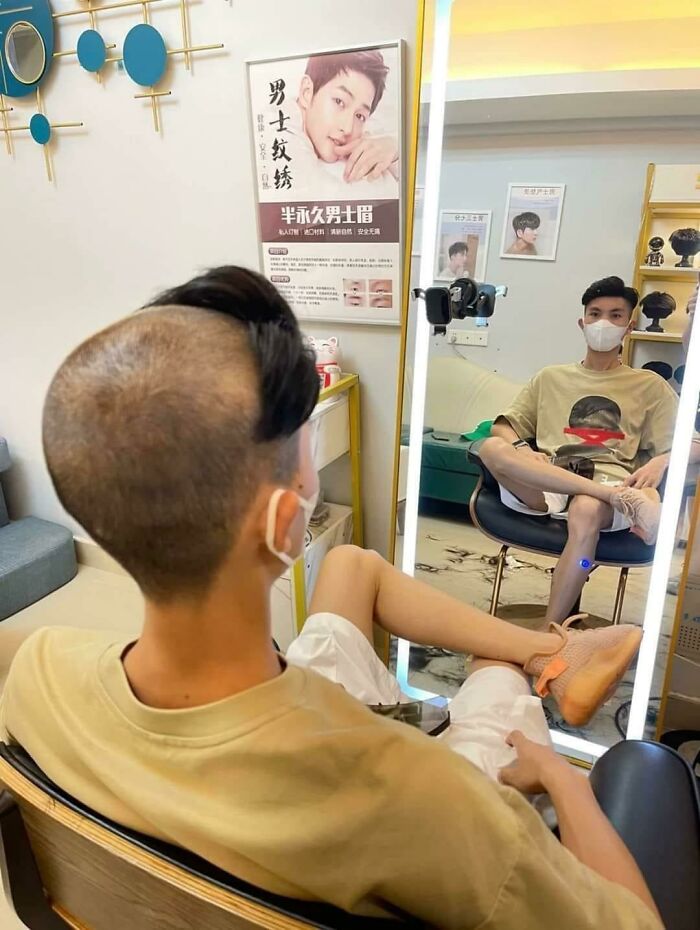 Man with a unique haircut sitting in front of a mirror in a hair salon, capturing his reflection with a mobile phone.