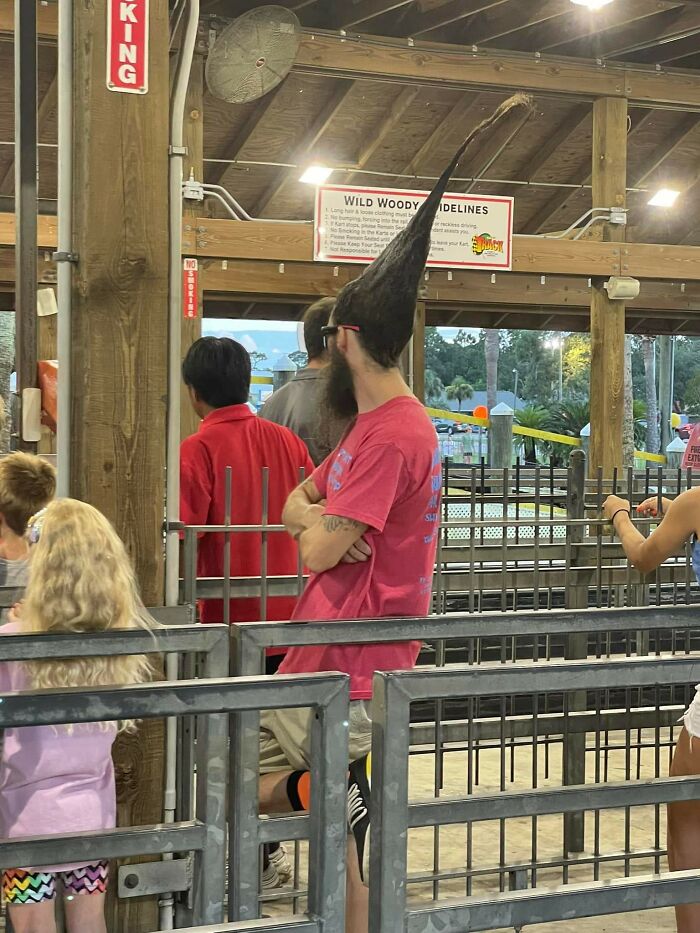 Man with a tall, unusual hairstyle stands in line at a theme park.