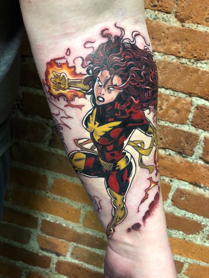 Dynamic Marvel tattoo featuring a superhero with fiery powers on a forearm against a brick background.
