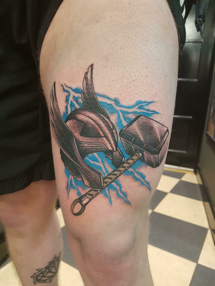 Marvel tattoo of a helmet and hammer with blue lightning on a person's thigh.