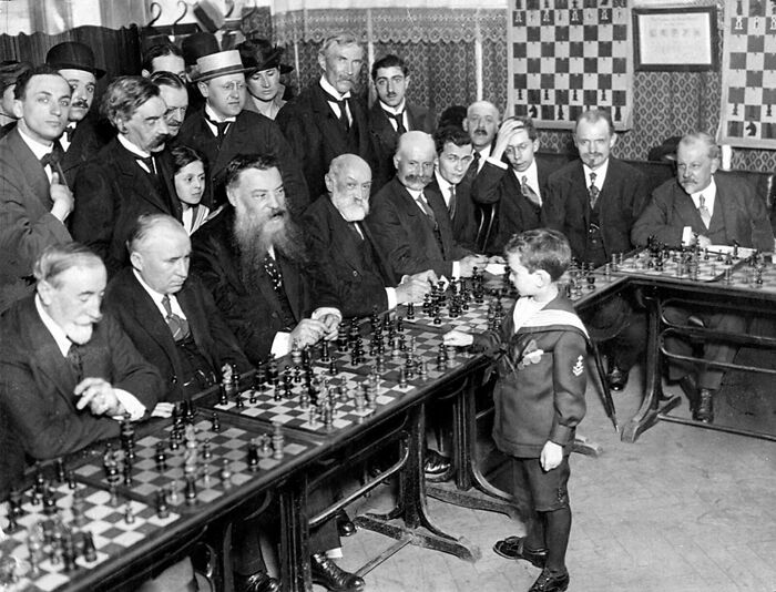 Young boy playing simultaneous chess against multiple older men in a historic event from around a century ago.