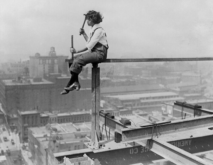 Young construction worker sitting on steel beam high above city, illustrating big historical events from a century ago.