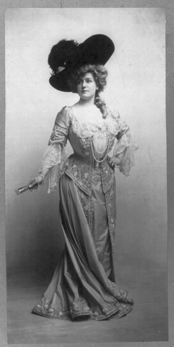 Victorian era woman in elaborate dress and large hat, representing big historical events from around a century ago.