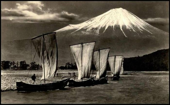 Vintage black and white photo of traditional sailboats near a shore with a snowy mountain in the background, historical events theme.