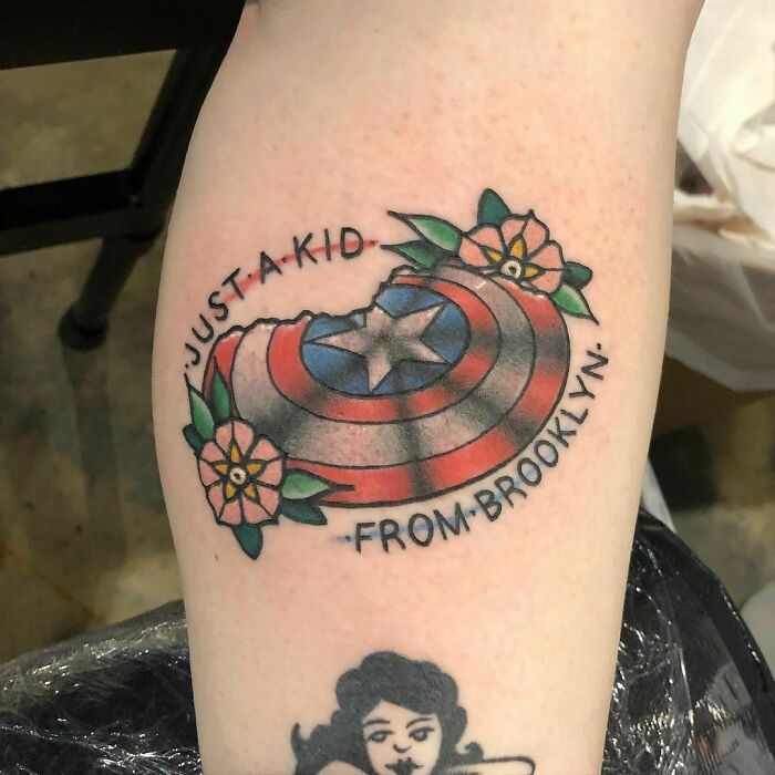 Marvel tattoo of Captain America's shield with floral design and text "Just a kid from Brooklyn" on arm.