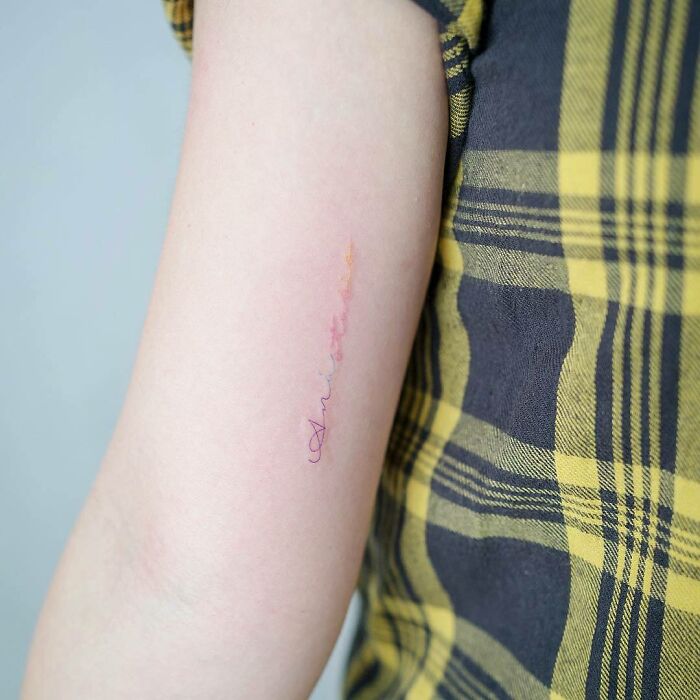 Minimalist word tattoo on arm with a plaid yellow shirt, using delicate colors.