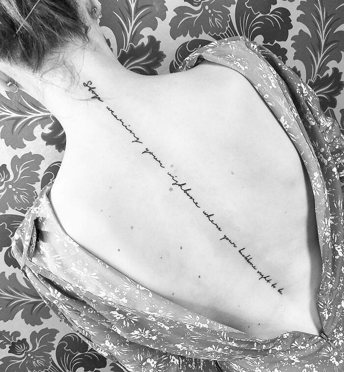 Elegant word tattoo in cursive script on a person's back, conveying creative inkspiration against a patterned wallpaper backdrop.