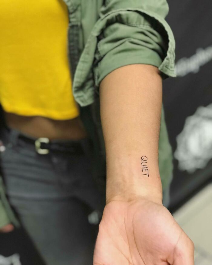 Word tattoo "quiet" on a forearm, wearing a yellow shirt and green jacket.
