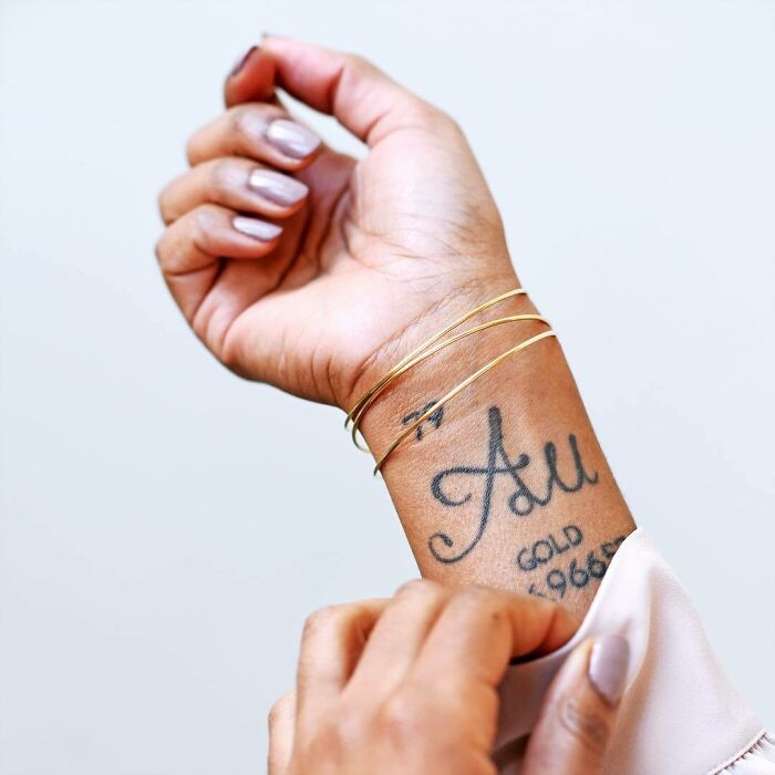 Close-up of a wrist with a creative word tattoo, featuring "Au" and "Gold" in elegant script, accessorized with a thin gold bracelet.