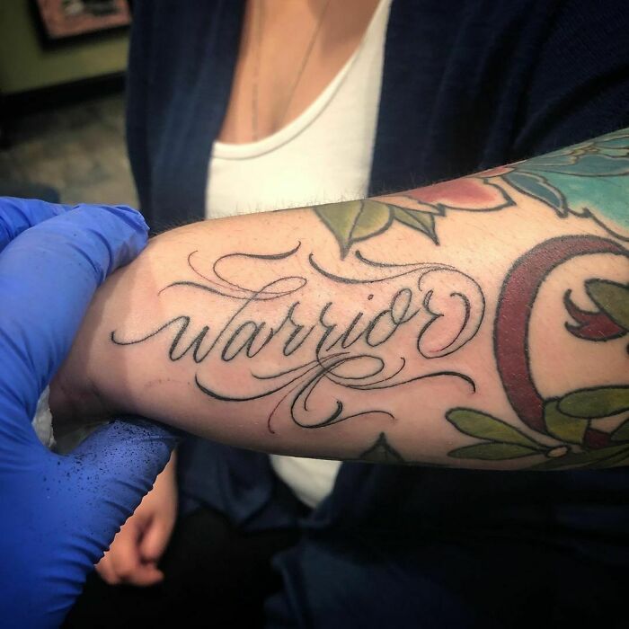 Arm tattoo with word "warrior" in elegant script, surrounded by colorful floral designs.