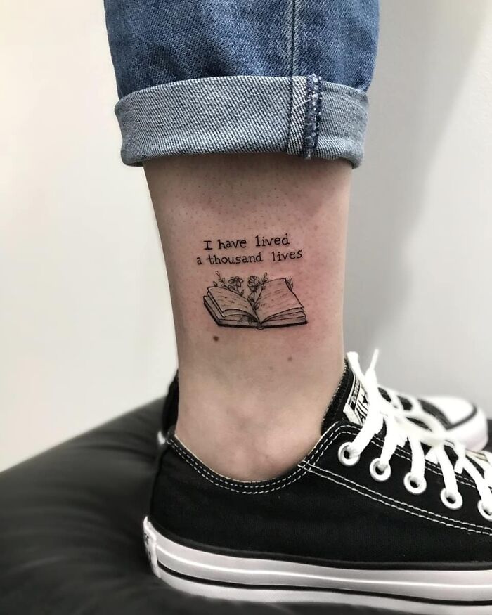 Creative word tattoo of an open book with flowers and text "I have lived a thousand lives" on a person's leg.