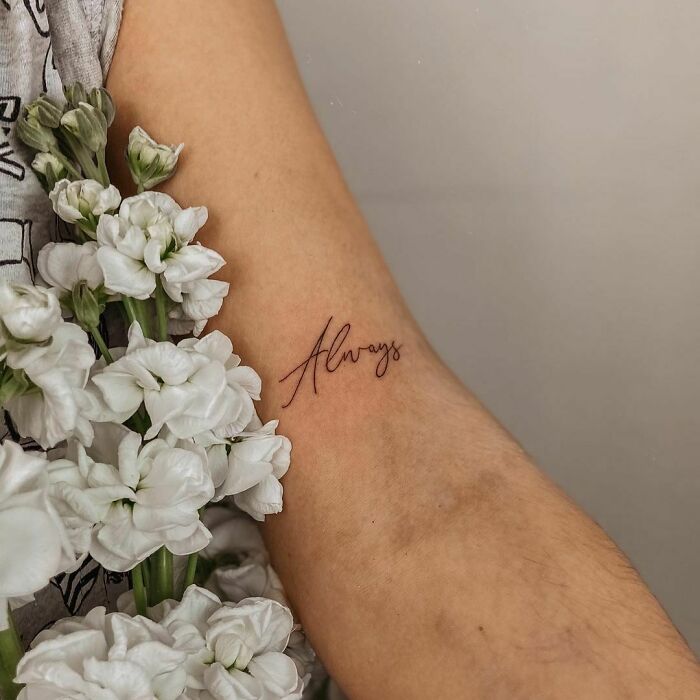 Delicate "Always" word tattoo on forearm surrounded by white flowers.
