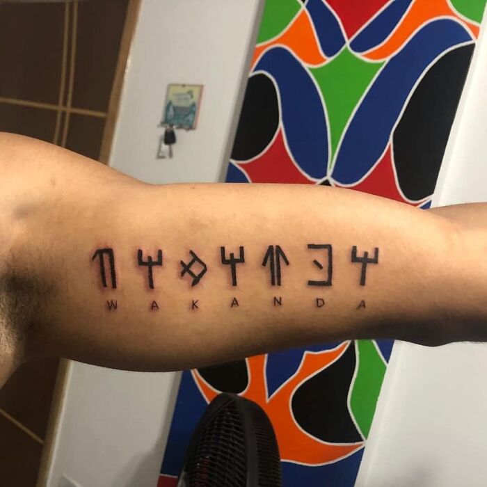 Arm tattoo featuring "WAKANDA" in stylized script, inspired by Marvel, against a colorful patterned background.