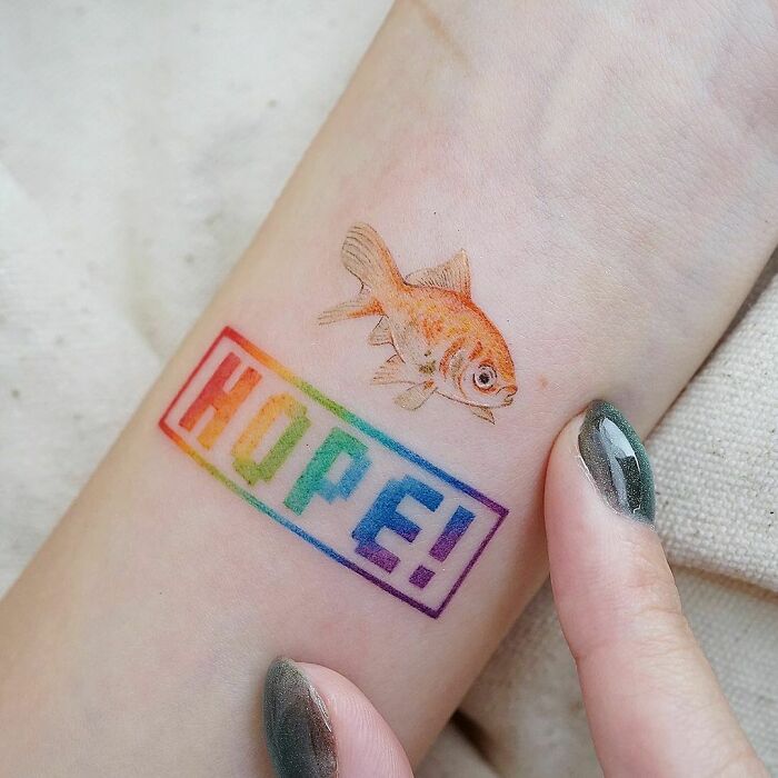 "Word tattoo with 'HOPE!' in rainbow colors and a fish depiction on forearm skin."