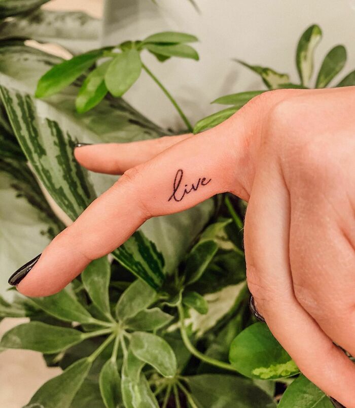 Word tattoo "live" on finger, surrounded by green leaves.