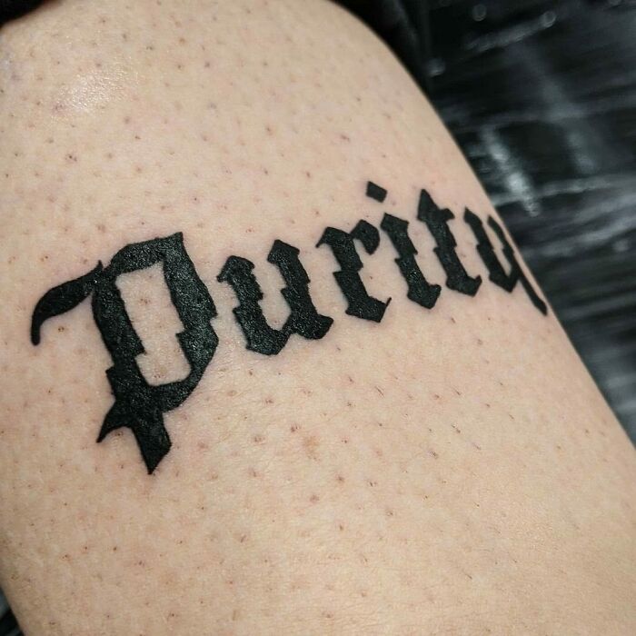 Word tattoo "Purity" in bold black lettering on skin.
