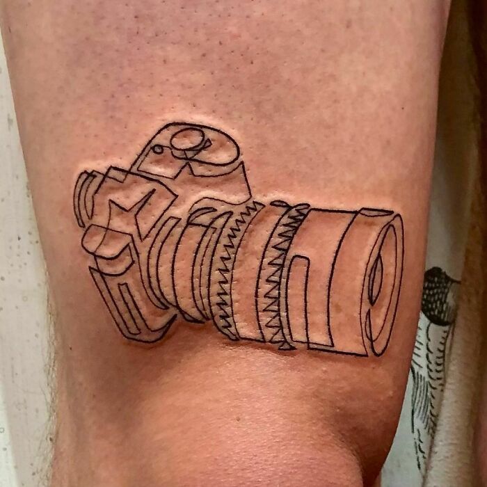Single-line tattoo of a camera on skin, showcasing fine-line perfection.