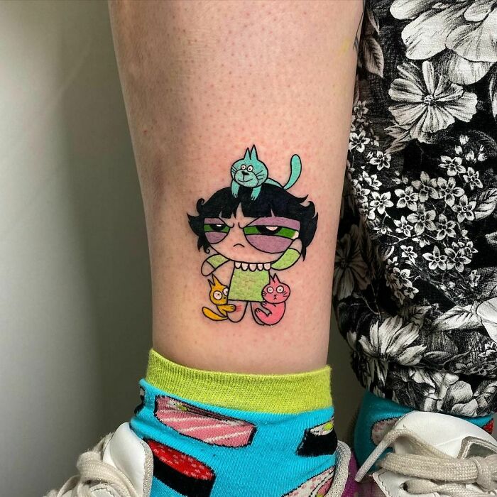Cartoon tattoo featuring a character with cats, inspired by animated shows, on a person's leg near floral leggings.