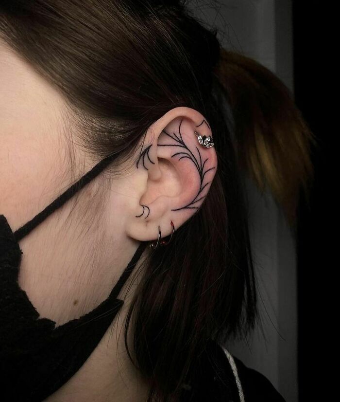 Delicate black linework ear tattoo with jewelry, worn by person with dark hair in a ponytail and mask.
