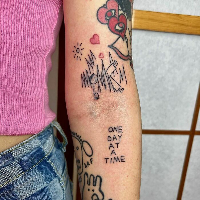 Black and red word tattoo ideas on an arm, featuring hearts and the phrase "one day at a time."