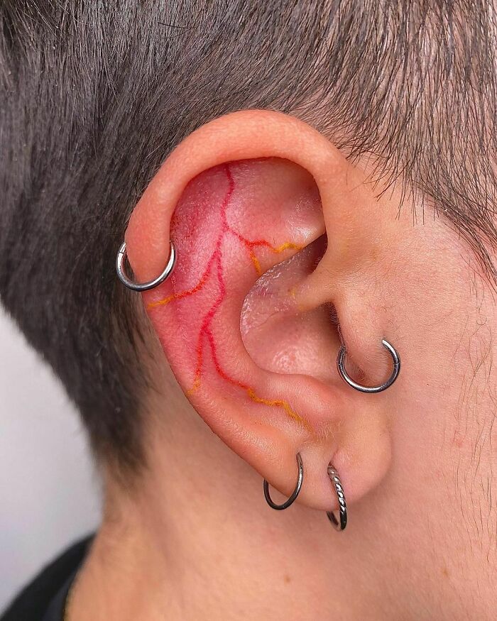 Ear tattoo with red and yellow lines, adorned with multiple silver hoop piercings, showcasing unique body art.