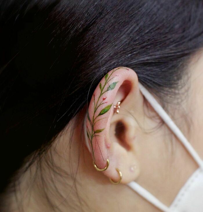 Delicate floral ear tattoo with green leaves and small pink flowers, complemented by gold hoop earrings.