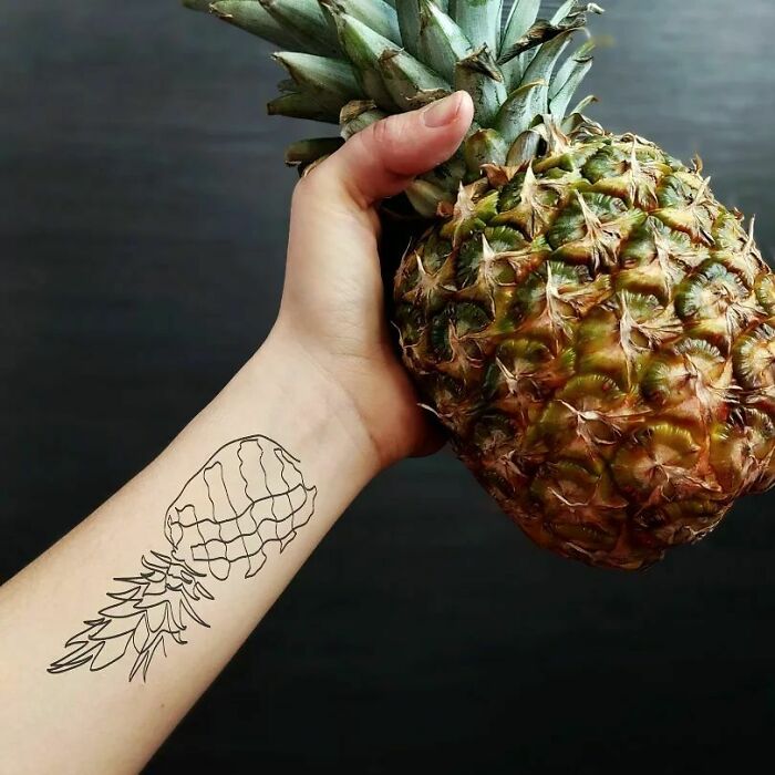 Arm with fine-line pineapple tattoo holding a real pineapple, showcasing single-line tattoo artistry.