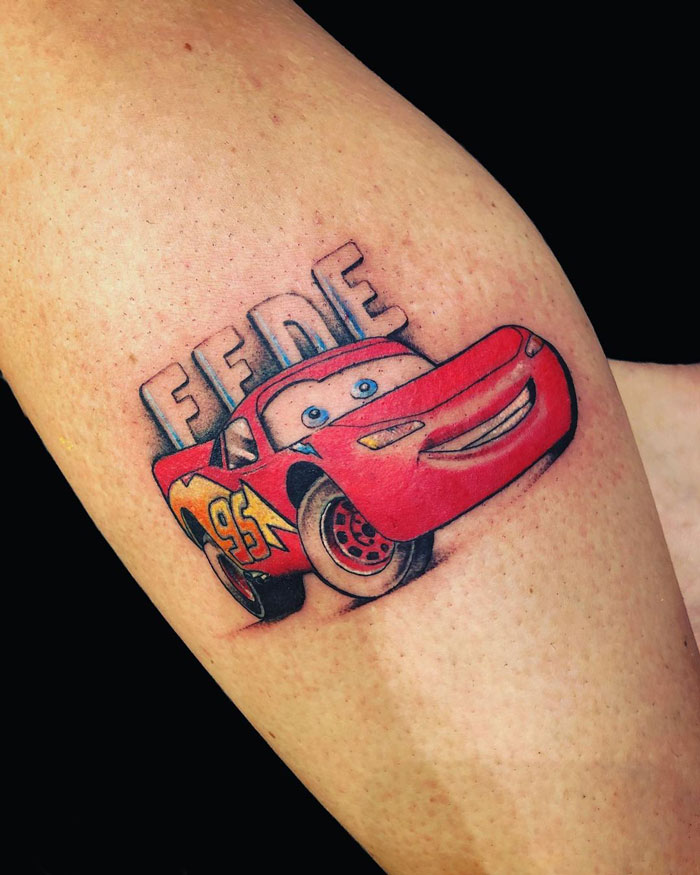 Cartoon tattoo featuring a smiling red race car Lightning Mcqueen with the text "FEDE" above it on a person's arm.