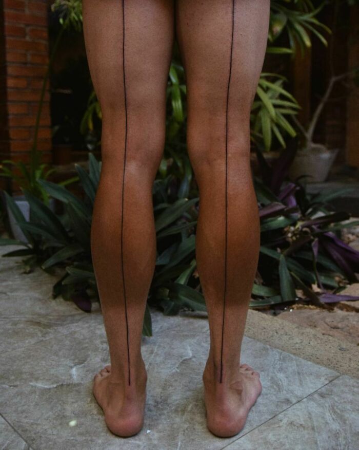 Bare legs with vertical single-line tattoos on calves, surrounded by plants.