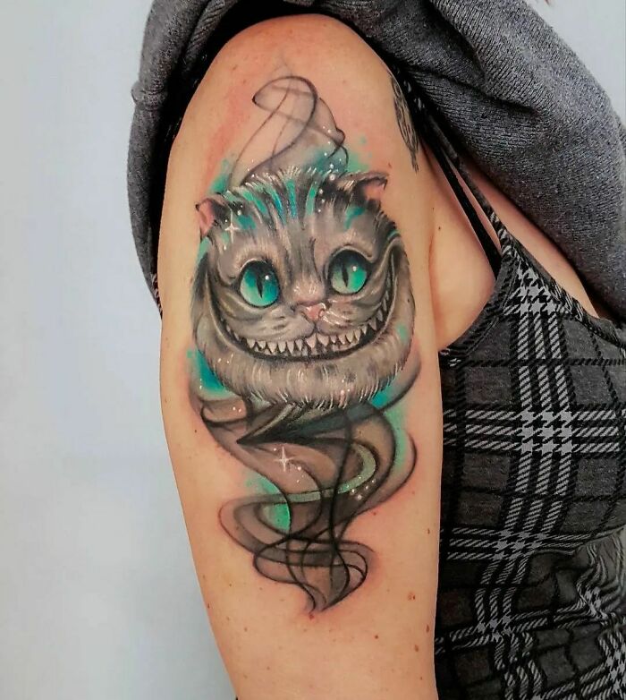 Cartoon tattoo of Cheshire Cat with bright colors on a person's upper arm, showcasing nostalgia and vivid artistry.