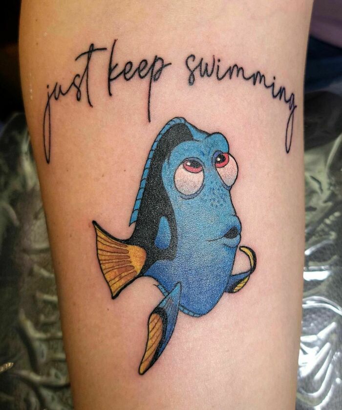 Cartoon tattoo of a blue fish with "just keep swimming" text, inspired by animated shows.