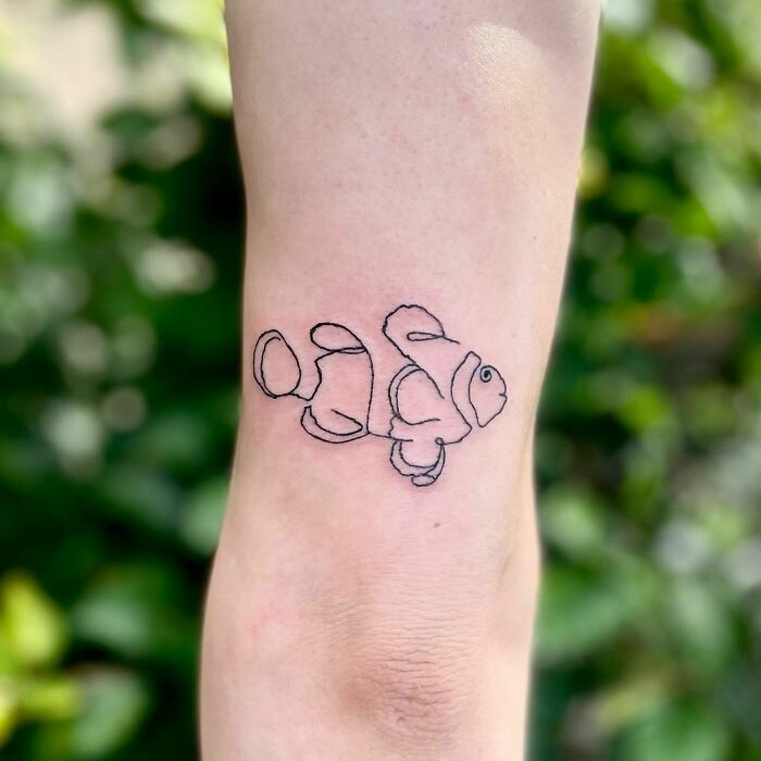 Single-line tattoo of a clownfish on an arm with a blurred green background.