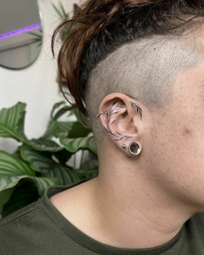 Shaved side hairstyle showcasing a subtle ear tattoo design with a feather pattern and piercings, set against a leafy background.
