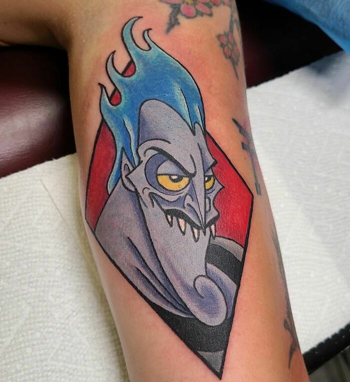 Cartoon tattoo featuring a blue-haired character Hades with a mischievous grin, inked on an arm.