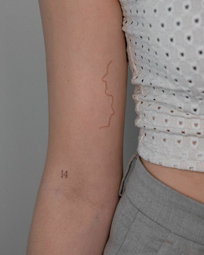 Unique line tattoo on arm with minimalist design and small number 14, captured against a patterned white top.