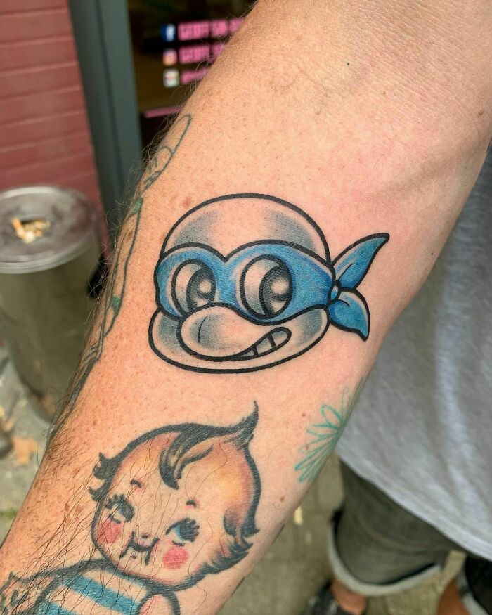 Cartoon tattoo of a blue-masked character on an arm, inspired by animated shows.