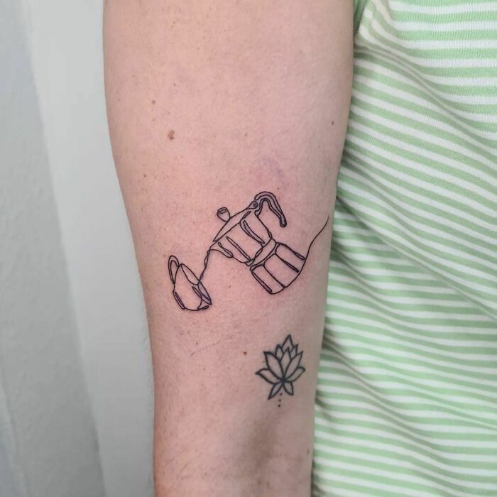 Single-line tattoo of a coffee maker on an arm, next to a small lotus design, showcasing minimalist ink artistry.
