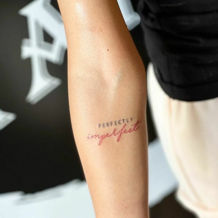 Arm with word tattoos "Perfectly imperfect" in black and red ink, showcasing meaningful tattoo art.
